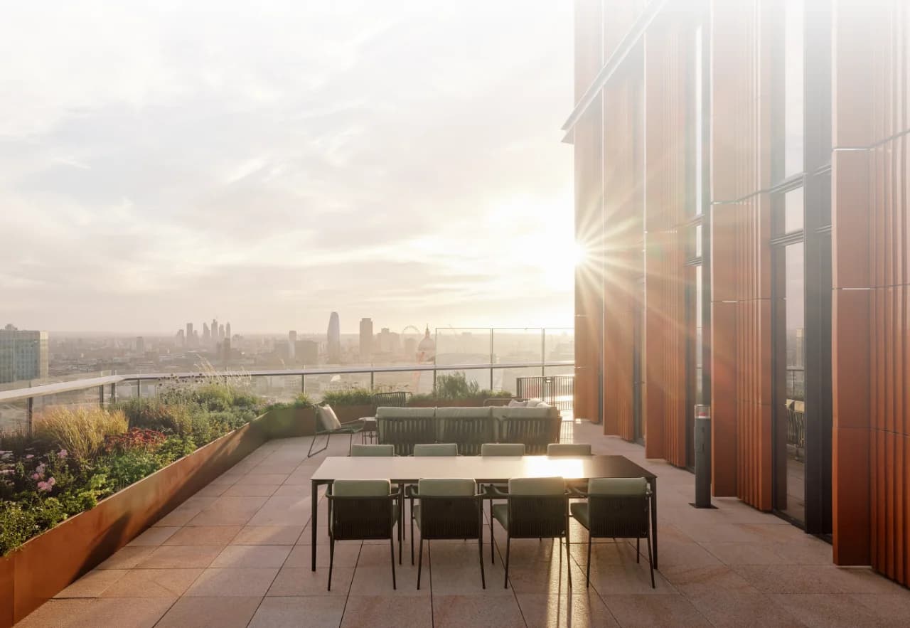 Rooftop terrace at sunset with outdoor dining table and lounge seating overlooking the London skyline, sunlight flaring beside a modern building facade.
